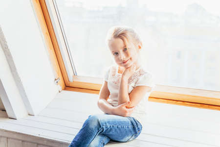 Stay Home Stay Safe. Little cute sweet smiling girl in jeans and white T-shirt sitting on window sill in bright light living room at home indoors. Childhood schoolchildren youth relax conceptの写真素材