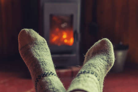 Feet legs in winter clothes wool socks at fireplace background. Woman sitting at home on winter or autumn evening relaxing and warming up. Winter and cold weather concept. Hygge Christmas eveの写真素材