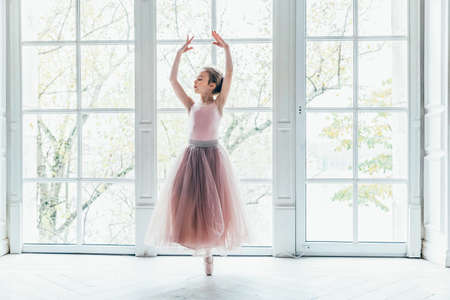 Young classical ballet dancer girl in dance class. Beautiful graceful ballerina practice ballet positions in pink tutu skirt near large window in white light hallの写真素材