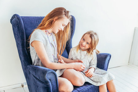 Two happy kids sitting on cozy blue chair relaxing playing in white living room indoors. Little girl playing with teenage girl showing her love care. Sisters having fun at homeの写真素材