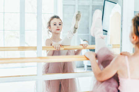 Young classical ballet dancer girl in dance class. Beautiful graceful ballerina practice ballet positions in pink tutu skirt near large mirror in white light hallの写真素材