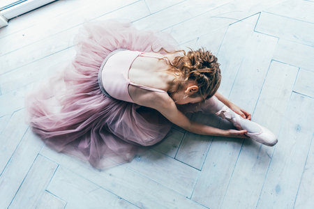 Young classical ballet dancer girl in dance class. Beautiful graceful ballerina in pink tutu skirt puts on pointe shoes near large window in white light hallの写真素材