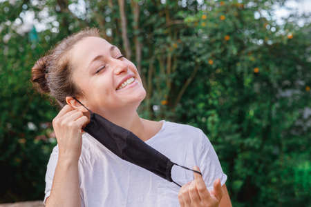 Happy positive girl takes off protective medical mask from face outdoors. Young woman removing mask smiling after vaccination.  Spring flowers pollen allergy.の写真素材