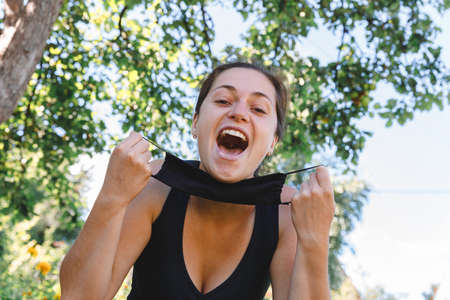 Happy positive girl takes off protective medical mask from face outdoors. Young woman removing mask smiling after vaccinationの写真素材