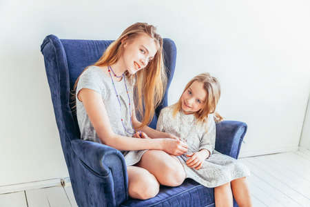 Two happy kids sitting on cozy blue chair relaxing playing in white living room indoors. Little girl playing with teenage girl showing her love care. Sisters having fun at homeの写真素材