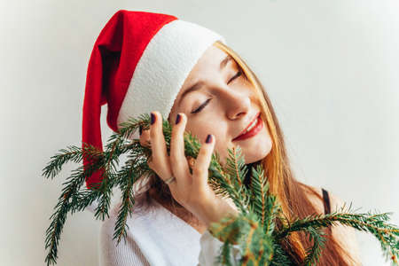 Beautiful girl in red Santa Claus hat holding fir tree branch isolated on white background looking happy and excited. Young woman portrait, true emotions. Happy Christmas and New Year holidaysの写真素材