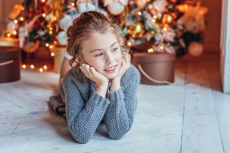 Little girl with gift box near Christmas tree on Christmas eve at home. Young kid in light bedroom with winter decoration Happy family at home Christmas New Year december time for celebration conceptの写真素材