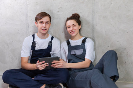 House DIY renovation concept. Young happy couple sitting on floor looking home repair plan in tablet indoors. Woman showing to man plan interior design online. Couple in new home during repair worksの写真素材