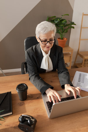 Confident stylish european middle aged senior woman using laptop at workplace. Stylish older mature 60s gray haired lady businesswoman sitting at office table. Boss leader teacher professional workerの写真素材