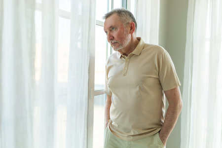 Sad tired lonely disappointed older senior man at home alone. Unhappy grandfather looking sadly outside window experiencing grief relative death bad news. Stressed elder man suffering from lonelinessの写真素材