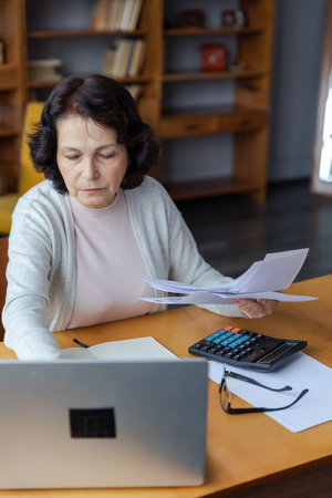 Middle aged senior woman sit with laptop and paper document. Pensive older mature lady reading paper bill pay online at home managing bank finances calculating taxes planning loan debt pension paymentの写真素材