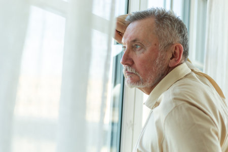 Sad tired lonely disappointed older senior man at home alone. Unhappy grandfather looking sadly outside window experiencing grief relative death bad news. Stressed elder man suffering from lonelinessの写真素材