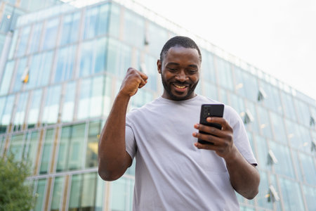 Happy African American man euphoric winner with smartphone on street in city. Person guy looking at cell phone reading great news getting good result winning online bid feeling amazed. Winning gestureの写真素材