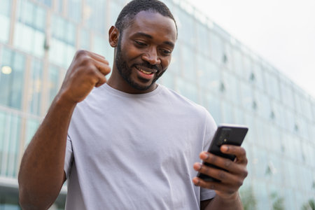Happy African American man euphoric winner with smartphone on street in city. Person guy looking at cell phone reading great news getting good result winning online bid feeling amazed. Winning gestureの写真素材