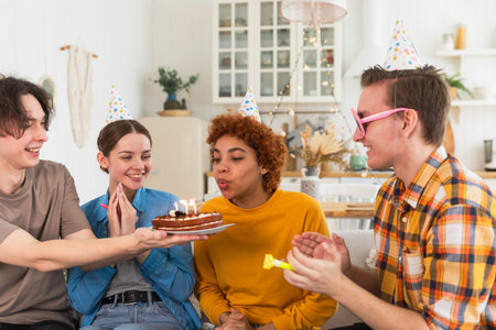 Make a wish. Woman wearing party cap blowing out burning candles on birthday cake. Happy Birthday party. Group of friends wishes girl happy birthday. People celebrating birthday with party at homeの写真素材