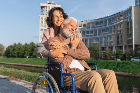 Old man in wheelchair walking with caregiver senior woman on road in park. Elderly family couple woman supporting embracing paralyzed man in chair for people with disability outdoor. Rehabilitationの写真素材
