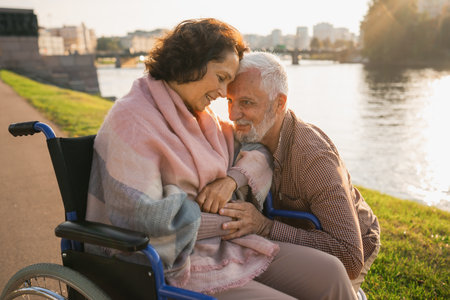 Senior woman in wheelchair walking with caregiver old man on road in park. Elderly family couple man supporting embracing woman in chair for people with disability outdoor. Rehabilitationの写真素材