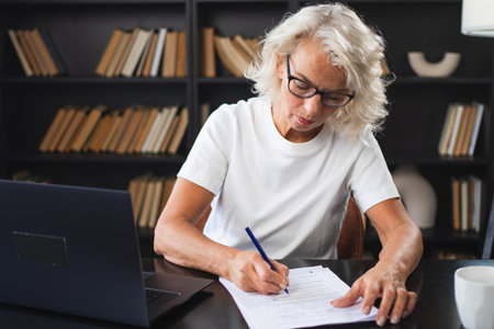 Middle aged senior woman using laptop computer writing notes at workplace. Focused mature old businesswoman work at office making written records doing online work. Mature boss professional workerの写真素材
