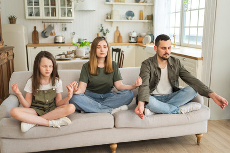 Yoga mindfulness meditation emotional wellbeing. Happy family practicing yoga at home Mother father daughter sitting in lotus pose meditating relaxing on couch Family doing breathing practice togetherの写真素材