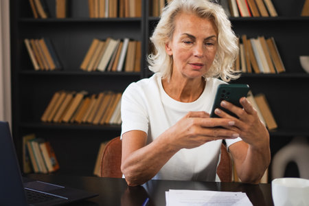 Confident stylish European middle aged senior woman using smartphone at workplace. Stylish older mature 60s lady businesswoman with cell phone in office. Boss leader using internet appsの写真素材