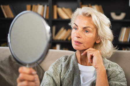 Love yourself. Beautiful old mature woman enjoying mirror reflection. Senior adult older lady looking at mirror confident happy on sofa at home. Woman doing daily morning beauty routine. Self loveの写真素材