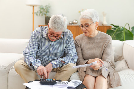 Middle aged senior couple sit with laptop and paper document. Older mature man woman reading paper bill pay online at home managing bank finances calculating taxes planning loan debt pension paymentの写真素材