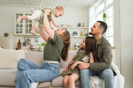 Happy family at home. Mother father two children daughters relaxing on sofa indoor. Mom dad parents baby girls kids relax playing having fun together. Family smiling laughing enjoying tender momentの写真素材