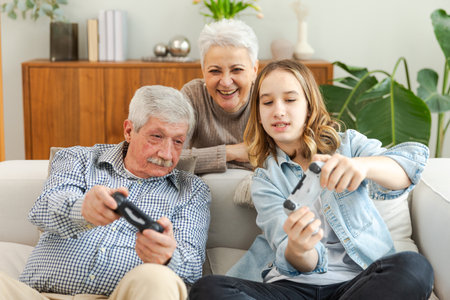 Happy family at home. Old senior man grandfather woman grandmother girl granddaughter playing video game with joysticks at home. Grandparent teenager two generations using gamepads for video gameの写真素材
