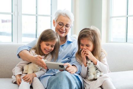 Happy family at home. Two little girls sisters twins grandmother enjoying time together watching video on phone. Good time at home. Grandma granddaughters child kids emotional bonding hugging together. Family generationsの写真素材