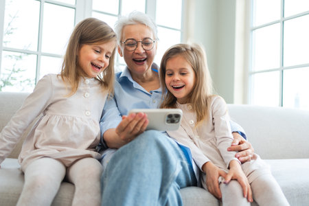Happy family at home. Two little girls sisters twins grandmother enjoying time together watching video on phone. Good time at home. Grandma granddaughters child kids emotional bonding hugging together. Family generationsの写真素材