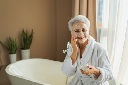 Mature adult old woman applying nourishing anti age face cream doing daily beauty routine. Senior woman with white mask or creme on skin gets facial treatment Skin care aging beauty skincare cosmeticsの写真素材