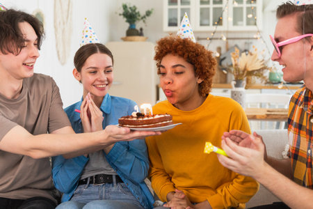Make a wish. Woman wearing party cap blowing out burning candles on birthday cake. Happy Birthday party. Group of friends wishes girl happy birthday. People celebrating birthday with party at homeの写真素材