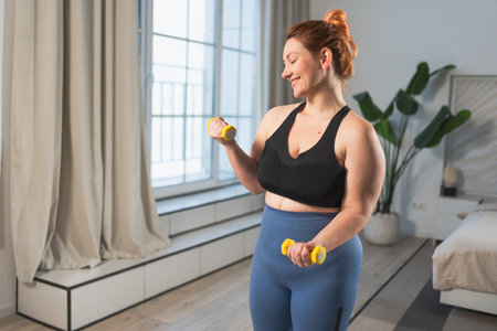 Fitness workout training. Woman picking up dumbbells. Plus size female doing sports exercise at home indoor. Girl in sportswear have workout weight training for weight loss. Sport and fitnessの写真素材