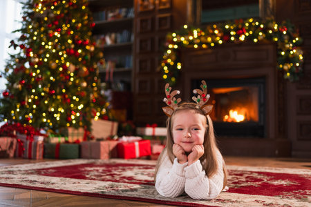 Merry Christmas. Little girl smiling near Christmas tree in classical dark interior. Young happy kid wearing deer horns in living room with fireplace Christmas tree gift boxes. Christmas eve at homeの写真素材