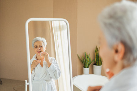 Love yourself. Beautiful old woman enjoying mirror reflection. Senior older lady spending morning in bathroom looking at mirror confident happy. Woman doing daily morning beauty routine. Self loveの写真素材
