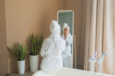 Mature adult old woman looking at mirror applying nourishing anti age face cream doing daily beauty routine. Senior woman with white mask creme on skin gets facial treatment Skin care aging skincareの写真素材