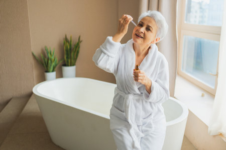 Woman applying natural drops cosmetics oil to face skin. Mature adult old woman applying nourishing anti age serum with pipette doing daily beauty routine. Senior woman gets facial treatmentの写真素材
