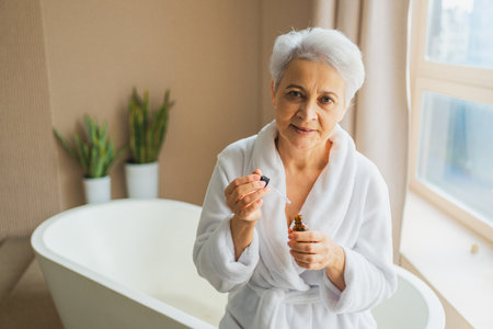 Woman applying natural drops cosmetics oil to face skin. Mature adult old woman applying nourishing anti age serum with pipette doing daily beauty routine. Senior woman gets facial treatmentの写真素材