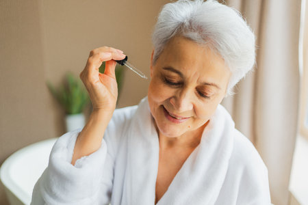 Woman applying natural drops cosmetics oil to face skin. Mature adult old woman applying nourishing anti age serum with pipette doing daily beauty routine. Senior woman gets kin scare facial treatmentの写真素材