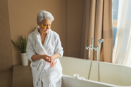 Mature adult old woman applying moisturizing body lotion on hands. Senior lady rubbing nourishing anti age cream doing morning daily skincare routine in bathroom. Beauty treatment spa body skin careの写真素材