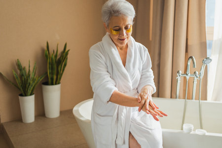 Mature adult old woman applying moisturizing body lotion on hands. Senior lady rubbing nourishing anti age cream doing morning daily skincare routine in bathroom. Beauty treatment spa body skin careの写真素材