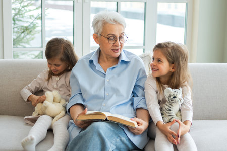 Happy family at home. Two girls sisters twins grandmother reading story book together. Good time at home. Grandma teaching granddaughters reading book fairytale telling story. Family generationsの写真素材