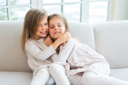 Happy family sisterly love. Two funny little twin sisters girls hugging with love care playing together at home indoors. Happy children lifestyle. Identical twin girls hugs. Girls friendship and youthの写真素材