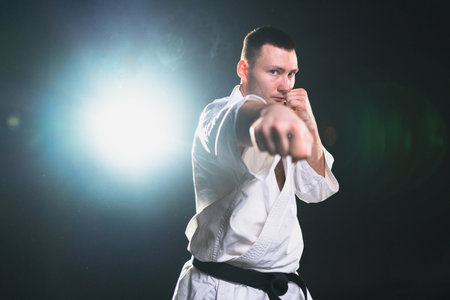 Strong man fighter in white kimono for karate martial arts with black belt preparing for fight training in black background. Man training moves punches kicks practicing shadow fight ready for sparringの写真素材