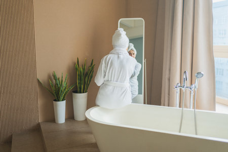 Mature adult old woman looking at mirror applying nourishing anti age face cream doing daily beauty routine. Senior woman with white mask creme on skin gets facial treatment Skin care aging skincareの写真素材