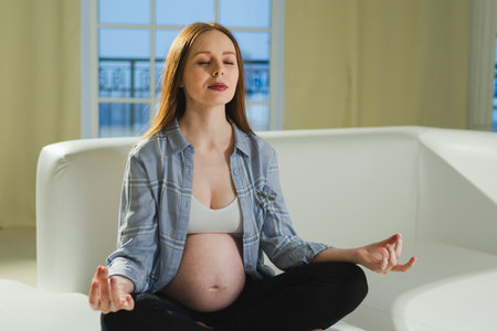 Pregnancy prenatal breathing. Pregnant woman with big belly sitting in lotus pose on sofa at home. Pregnant girl practicing yoga mindfulness meditation. Relaxation self care during pregnancyの写真素材