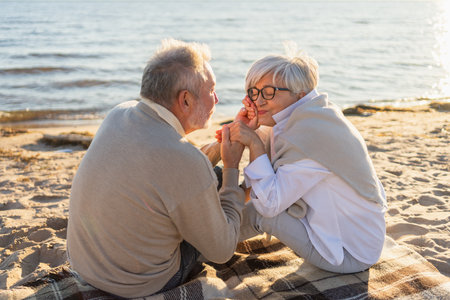Senior mature couple sitting on beach holding hands enjoying outdoor recreation. Old husband wife touching hands with tenderness love. Grandmother grandfather together. Family moment of loveの写真素材