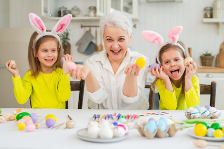 Happy Easter. Two little girls sisters twins grandmother painting eggs enjoying time together. Happy family grandma granddaughters child kids preparing for Easter. Spring Christian festival traditionの写真素材