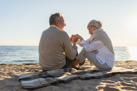 Senior mature couple sitting on beach holding hands enjoying outdoor recreation. Old husband wife touching hands with tenderness love. Grandmother grandfather together. Family moment of loveの写真素材