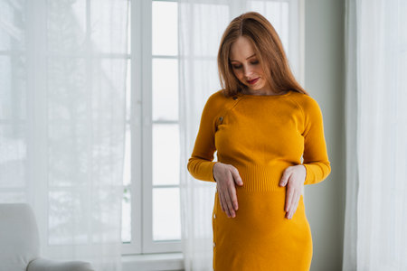 Pregnancy motherhood people expectation future. Pregnant woman with big belly standing near window at home. Girl hugging her tummy enjoying pregnancy. Maternity tenderness parenthood new life conceptの写真素材